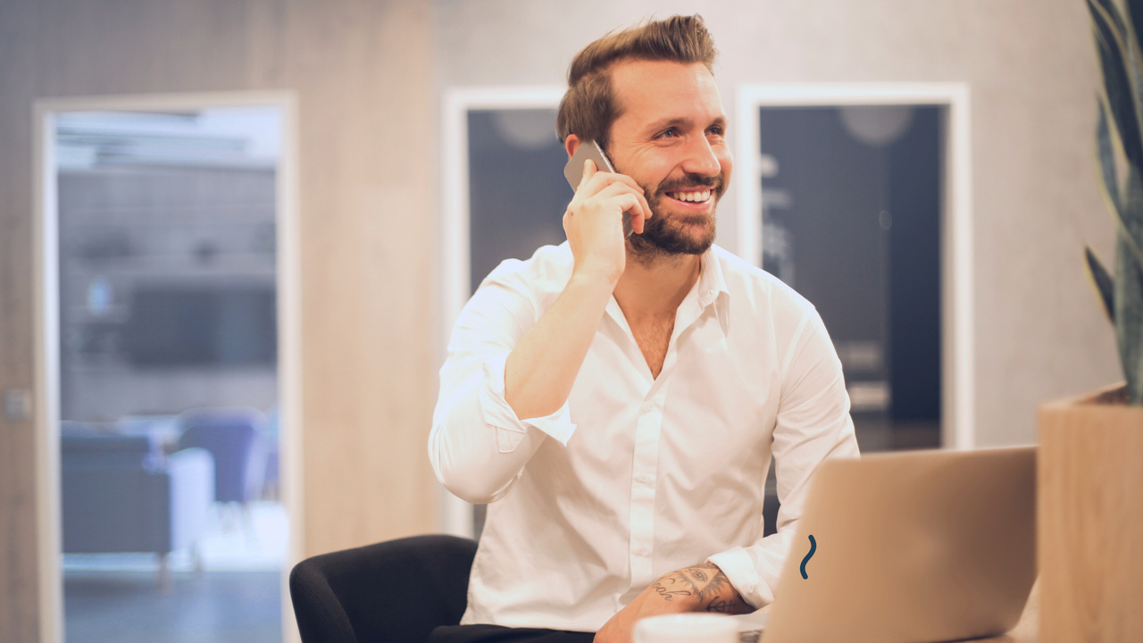 Profissional com telefone na orelha em frente ao computador. Imagem simboliza gerente comercial conversando com cliente.